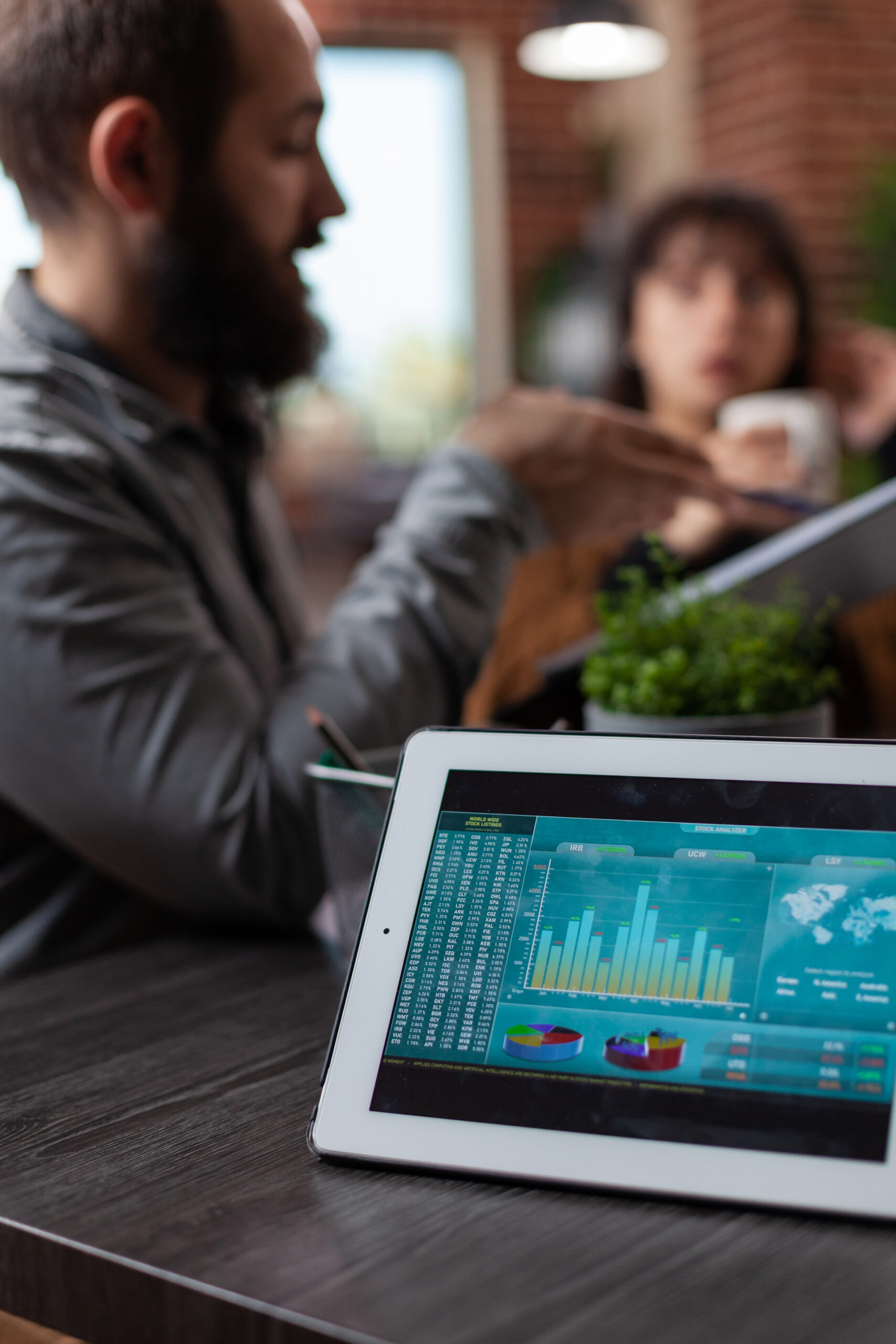 Tablet computer with company statistics on screen standing on table in startup office. Millennial multiethnic businesspeople discussing marketing strategy working at business collaboration